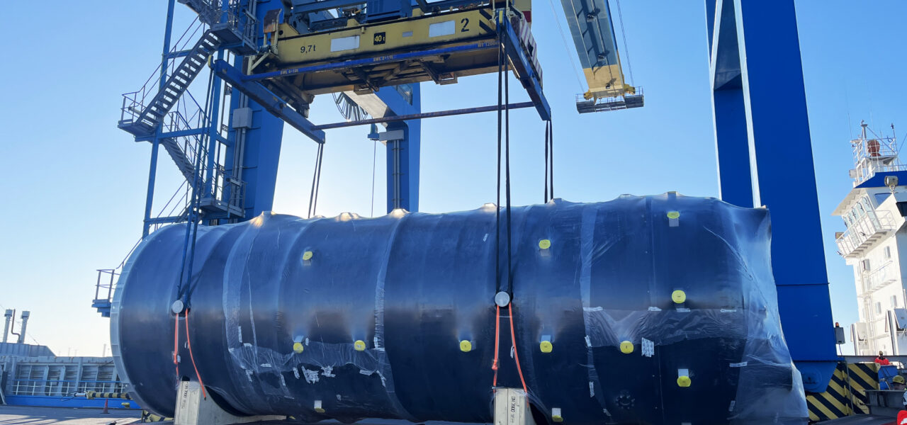 Large cylindrical industrial unit being lifted by crane onto trailer at port during project cargo handling