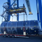 Large cylindrical industrial unit being lifted by crane onto trailer at port during project cargo handling
