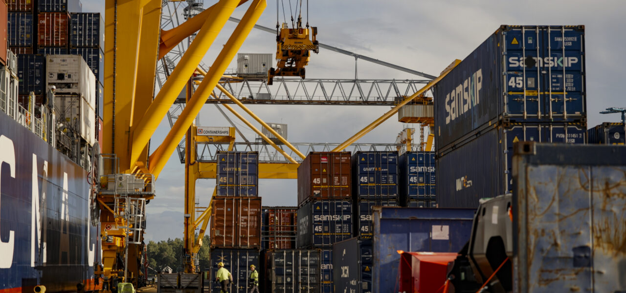 Container handling operations at a Finnish cargo terminal with gantry cranes, stacked shipping containers, and a container vessel alongside the quay.