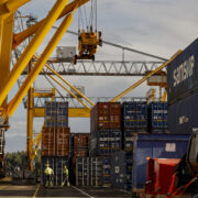 Container handling operations at a Finnish cargo terminal with gantry cranes, stacked shipping containers, and a container vessel alongside the quay.