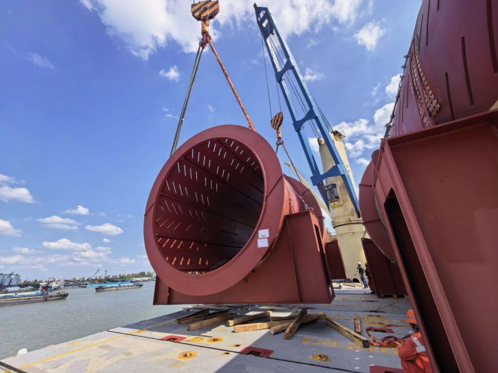 Heavy lift operation loading a large industrial drum onto a multipurpose vessel using ship and port cranes under clear skies on an Intra-Asia project cargo shipment.