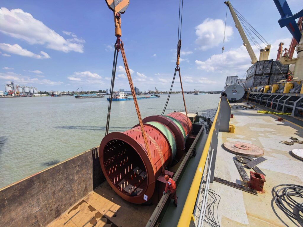 Large industrial process drum being lifted from a barge to a cargo vessel during a complex heavy-lift loading operation at an Asian port.