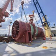 170-ton industrial drum secured on the deck of a heavy-lift vessel while cranes and stevedores complete cargo handling operations at the port.